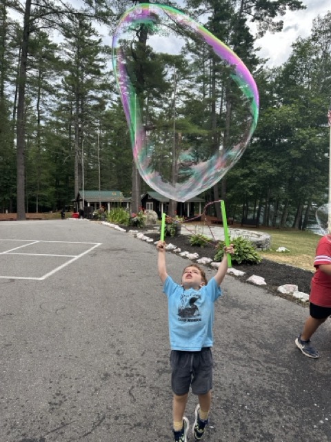 A Little Loon jumping under a giant rainbow bubble in front of the Wigwam Lodge