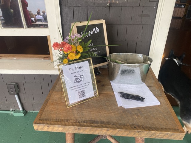 Welcome and registration table at a Camp Wigwam wedding with a 'Favors' chalkboard sign, fresh florals, a QR code photo share, and rustic details
