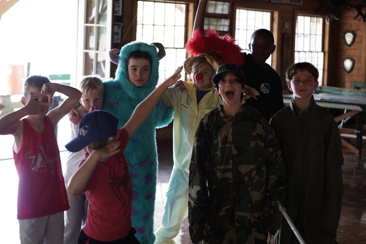 Group of campers in costumes — a Sully suit, clown, camo, and flight gear — posing inside the lodge