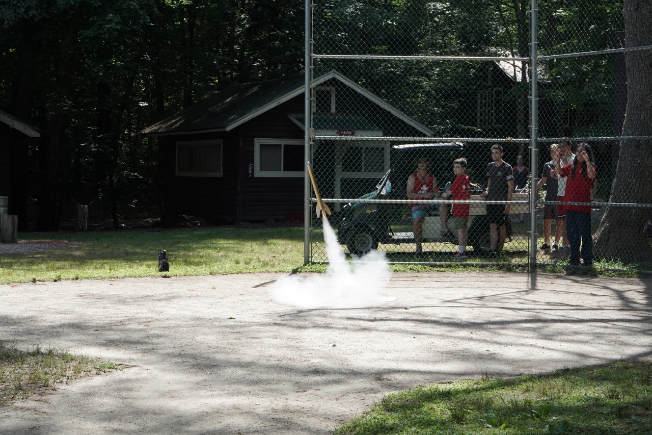 Model rocket lifting off from the launch pad as campers watch from behind a safety fence