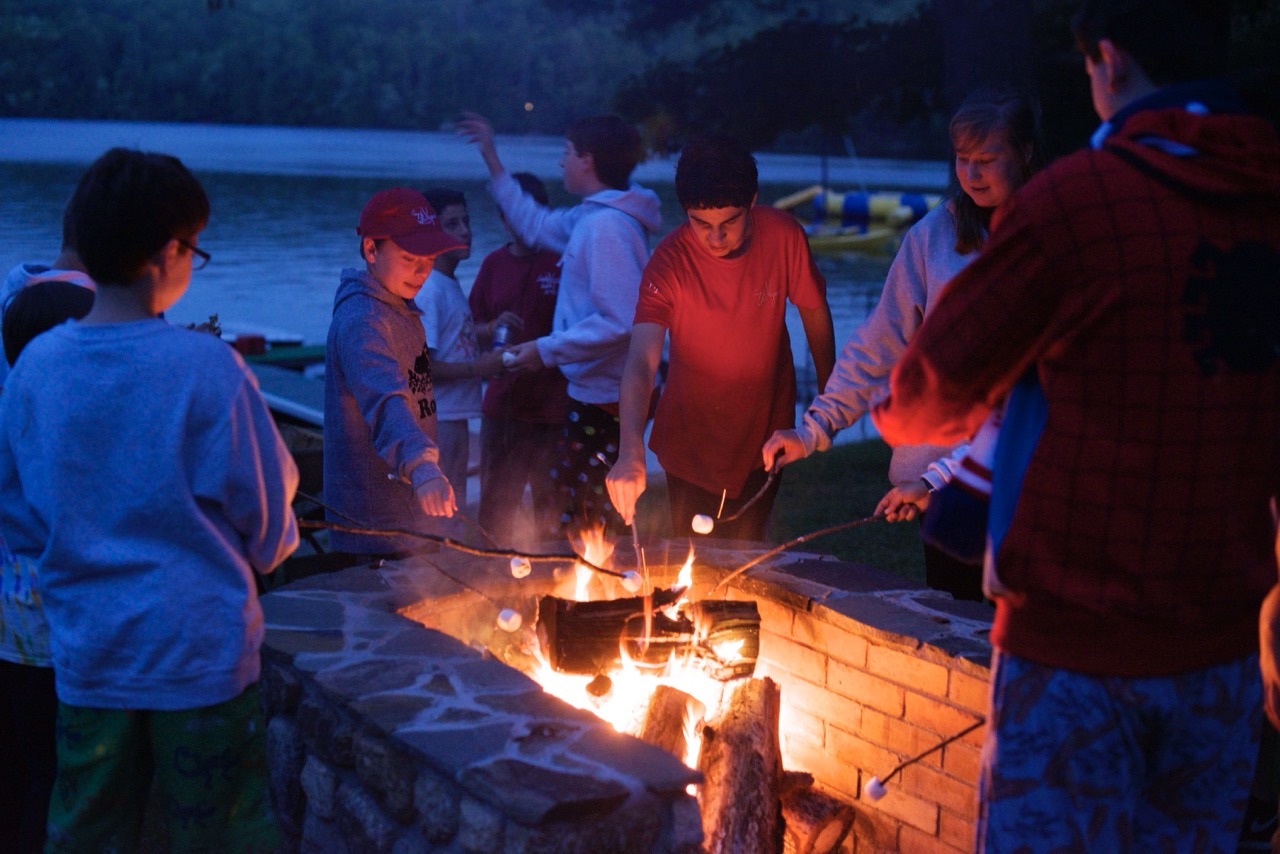 Campers and counselors gathered around the lakeside fire pit roasting marshmallows for s'mores at dusk