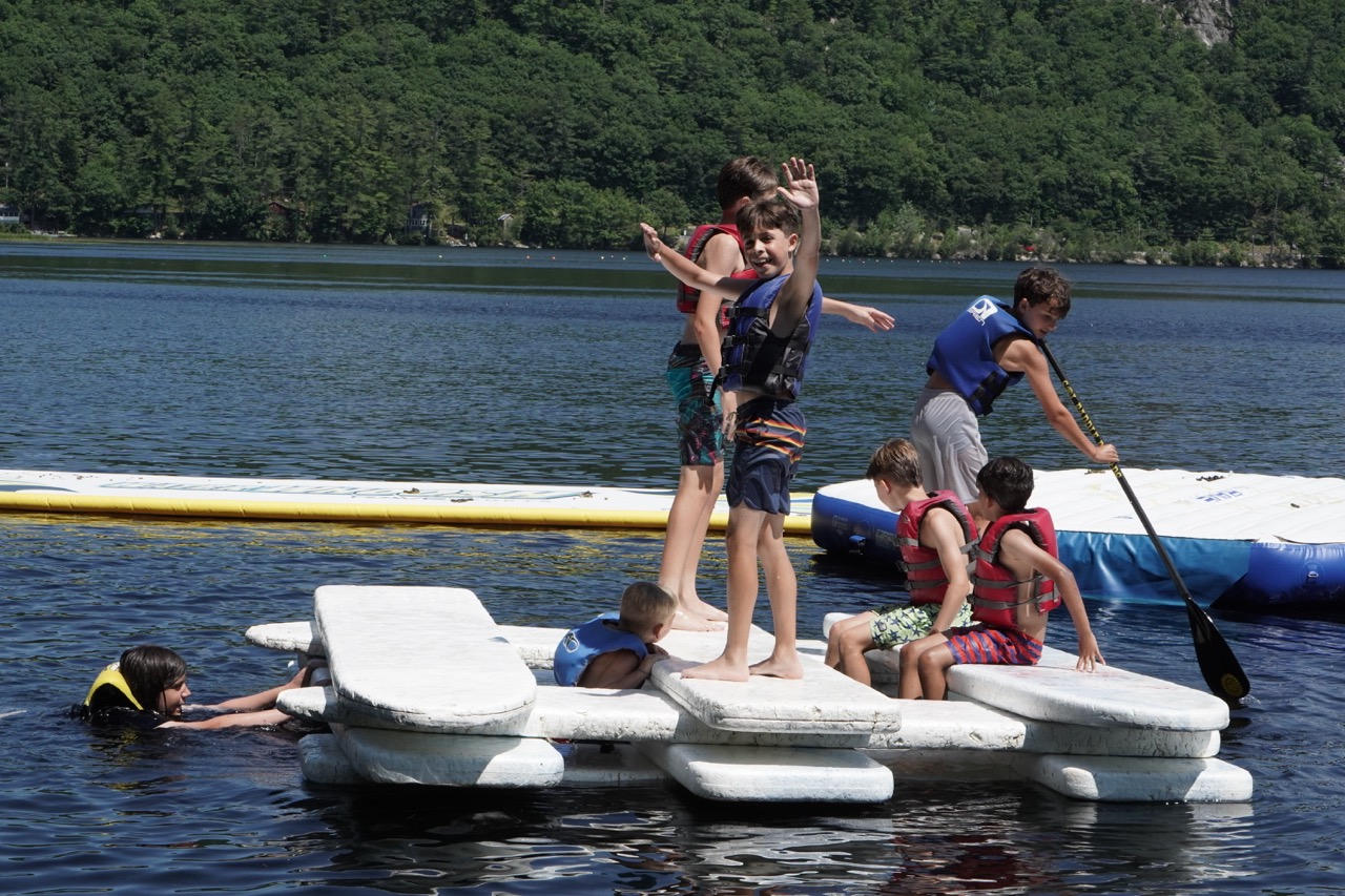 Group of campers in life jackets playing on floating mats and paddleboards at the Bear Lake waterfront